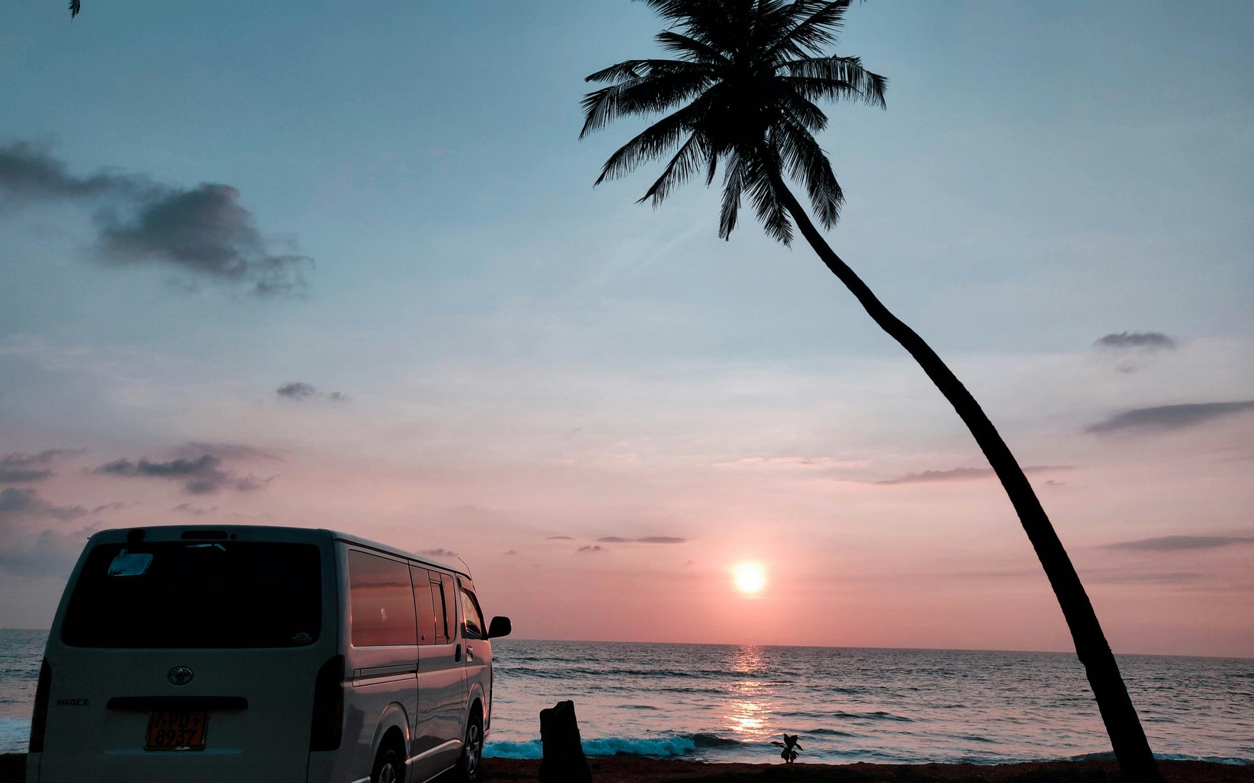 A chauffeur van parked on a Sri Lankan beach at sunset beneath a palm tree
